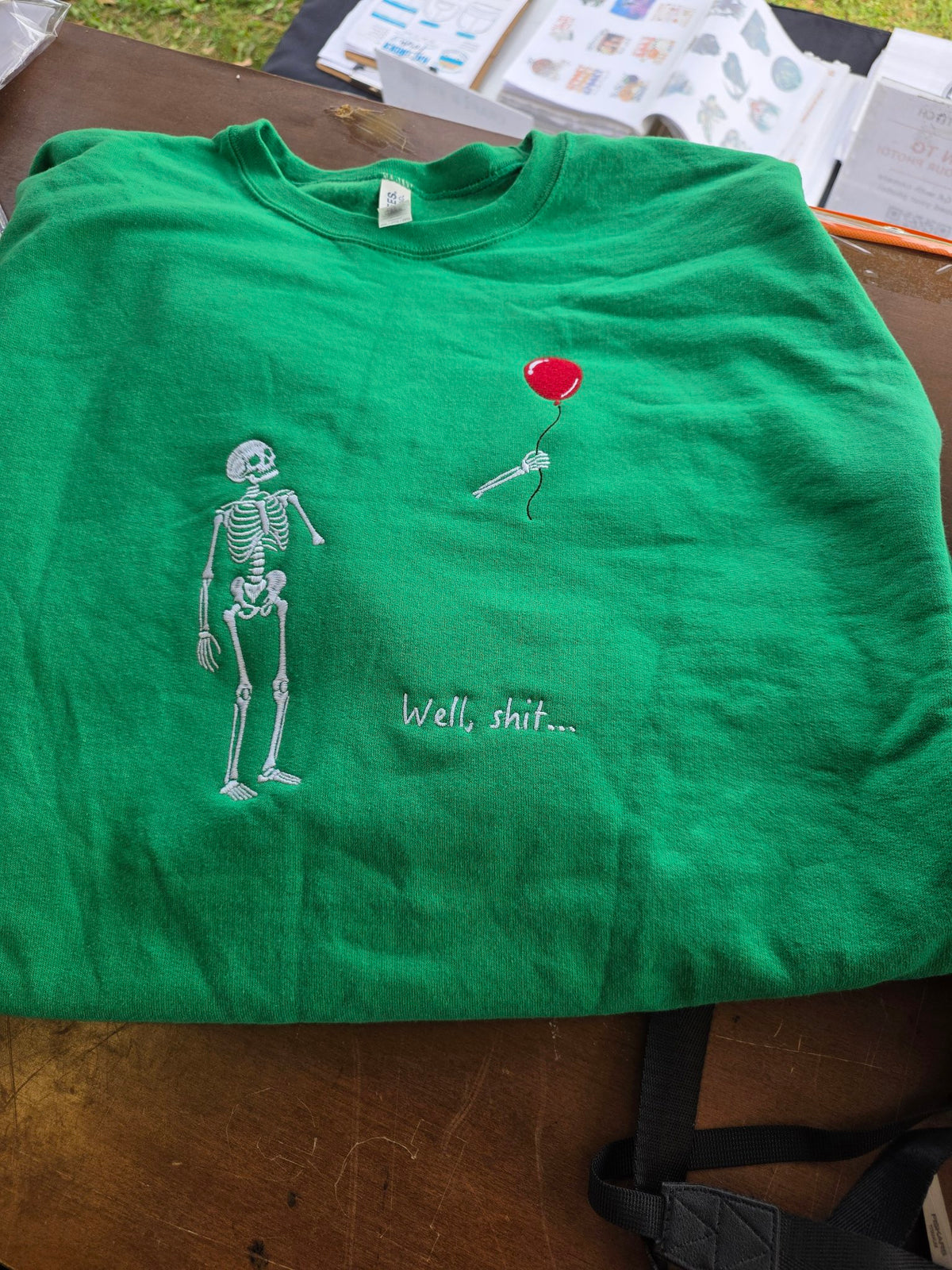 Embroidered skeleton holding red balloon with 'Well, shit...' text on green t-shirt